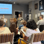 Instructor leading a Color Bootcamp workshop, speaking in front of a screen while students take notes and demo panels sit on easels.