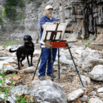 Duane Hada plein air paints on an Artefex all-in-panel set on an easel by a rocky creek with his dog at his side